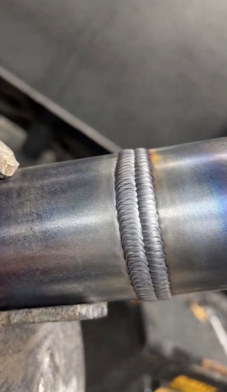 Two parallel stick weld beads with an even ripple pattern running around a carbon steel pipe