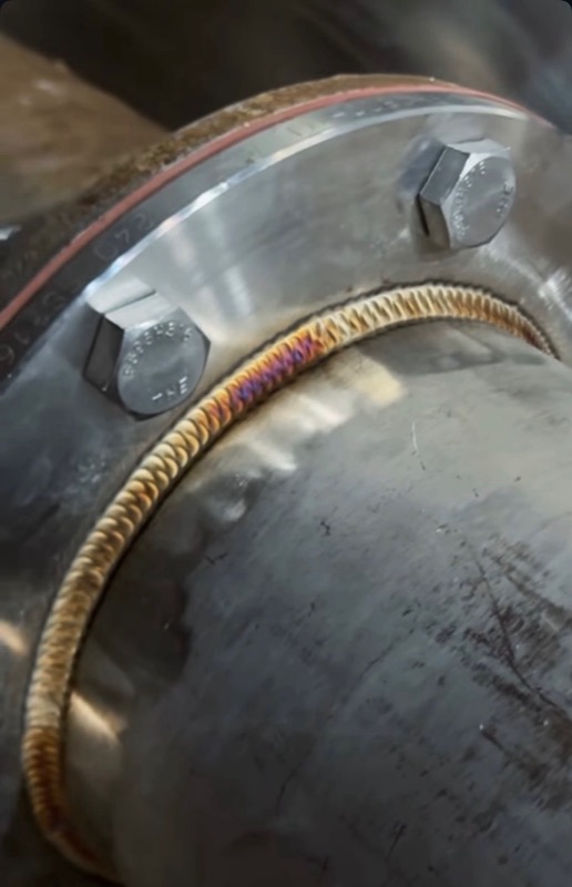 TIG weld bead with a gold heat tint curving around the radius of a bolted flange